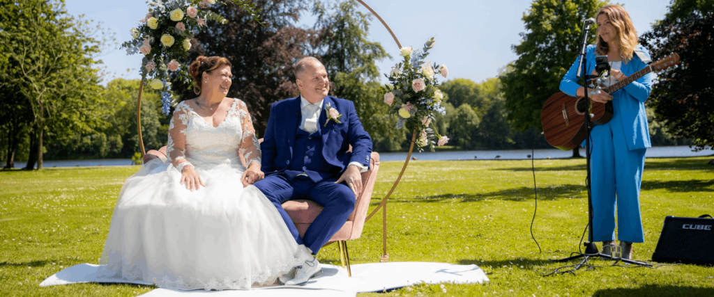 Je ziet een bruidspaar tijdens de trouwceremonie. Naast het bruidspaar staat een zangeres met akoestische gitaar.